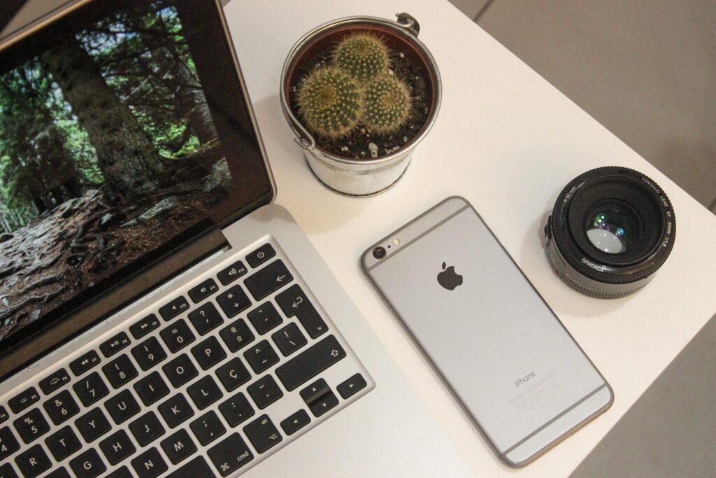 Desktop with MacBook, iPhone, small potted cactus, and camera lens.